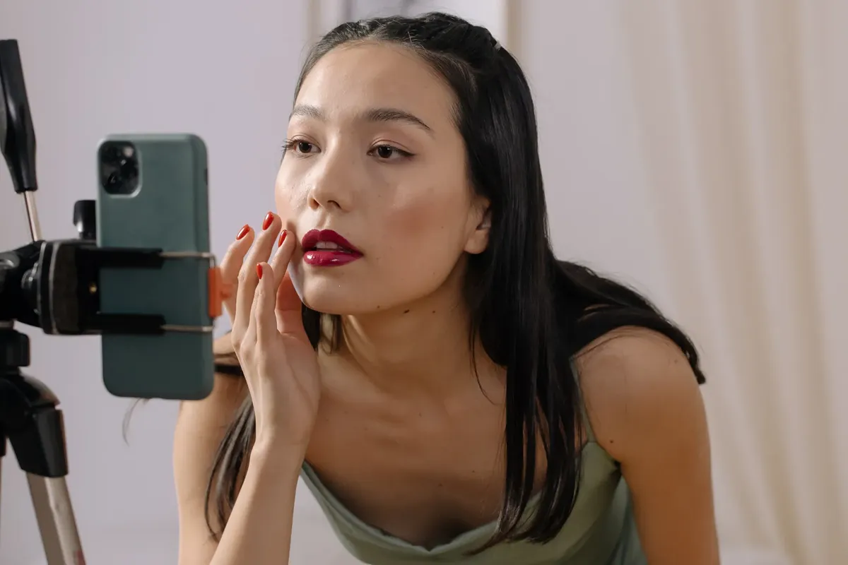 Asian woman applying red lipstick during a live streaming makeup vlog using a smartphone.